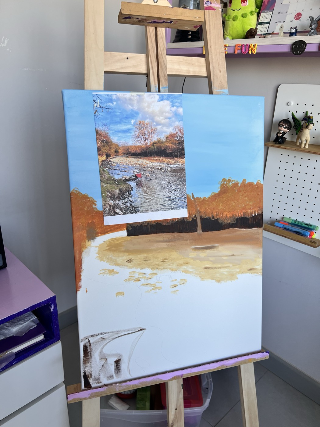 El cuadro de Toffy a medio pintar en el caballete: un río entre árboles otoñales, con la foto de referencia impresa pegada encima