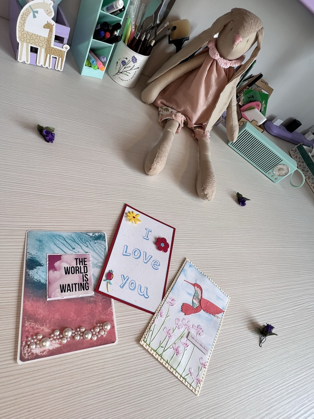 Tres tarjetas hechas a mano sobre la mesa de trabajo: una con fondo de acuarela azul y rosa con perlitas, una que dice I love you, y una con un colibrí entre flores